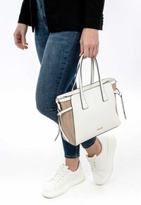 Sac fourre-tout blanc avec des panneaux latéraux texturés, des bordures noires, des double poignées et une fermeture éclair. Porté avec un jean bleu et des baskets blanches.