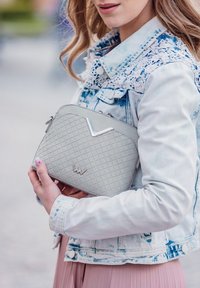 Šedá texturovaná kabelka s diamantovým vzorem, s kovovým akcentem a zipovým uzávěrem, držena proti světlé džínsové bundě.