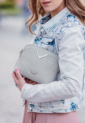 Šedá texturovaná kabelka s diamantovým vzorem, s kovovým akcentem a zipovým uzávěrem, držena proti světlé džínsové bundě.