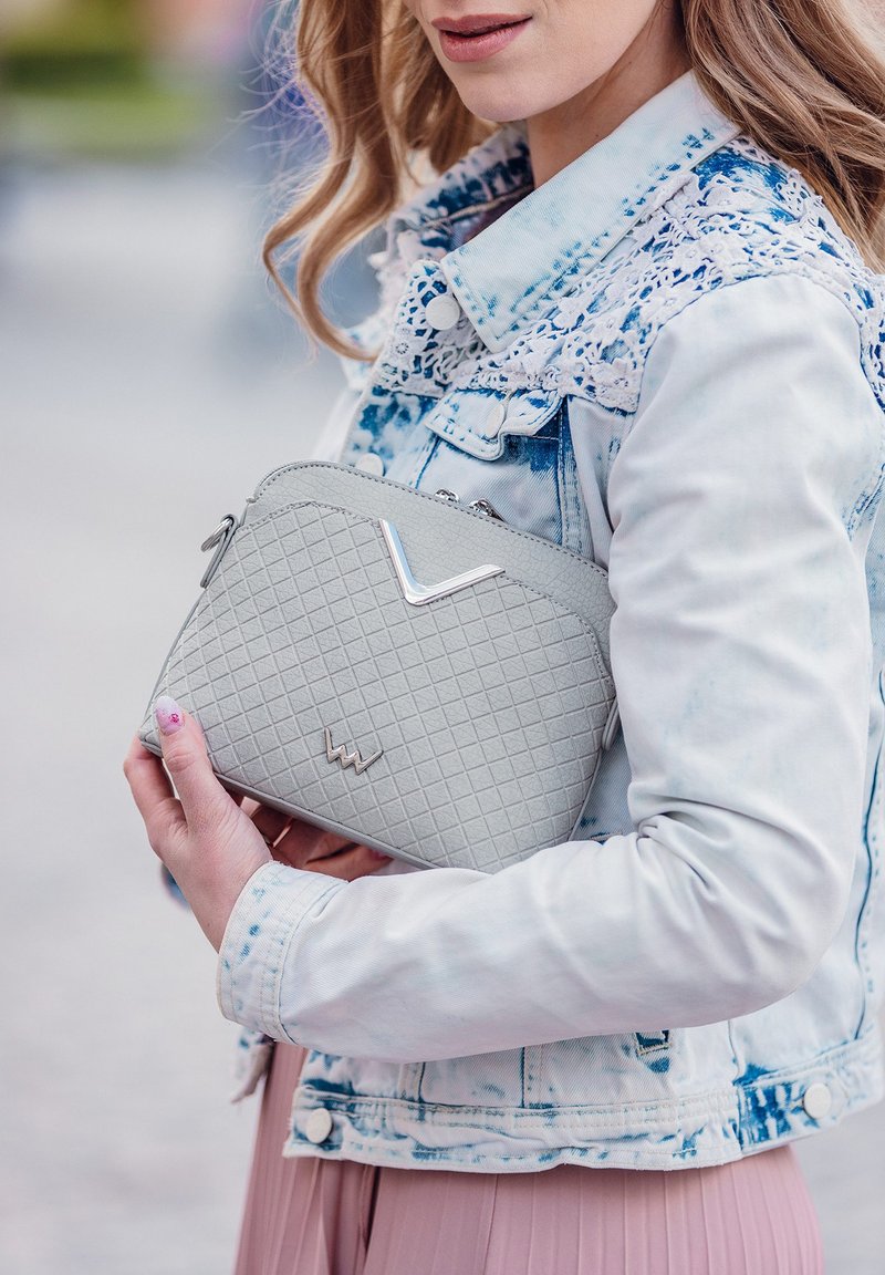 Šedá texturovaná kabelka s diamantovým vzorem, s kovovým akcentem a zipovým uzávěrem, držena proti světlé džínsové bundě.