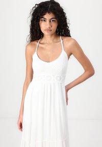 White maxi dress with a fitted crochet bodice and flowing skirt. Features thin straps and a textured, pleated fabric for a light appearance.