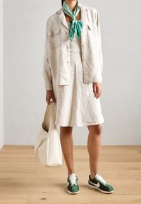 Giacca camicia beige in lino con due tasche, gonna corta abbinata, top bianco e sneakers verdi. Completato con una borsa tote bianca e un foulard verde.
