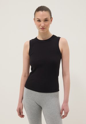 Frau mit zurückgebundenem Haar, die ein ärmelloses schwarzes geripptes Oberteil und hellgraue Leggings trägt, steht vor einem schlichten hellen Hintergrund.