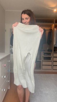 Vestido de malha branca canelada com uma fenda lateral, apresentando um tecido texturizado e uma bainha desfiada. O item está exposto numa sala com toques em madeira.