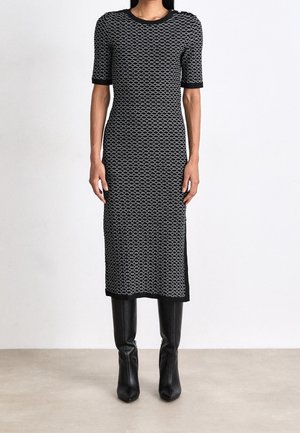 Mujer con vestido midi de patrón blanco y negro y mangas cortas, combinado con botas de cuero negras hasta la rodilla, de pie sobre un suelo neutro.