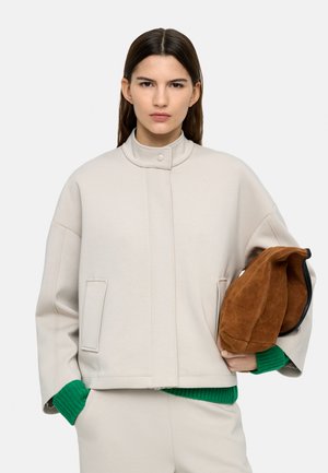 Kobieta o prostych brązowych włosach nosi jasnobeżową kurtkę i dopasowane spodnie, spod rękawów wystaje zielony sweter, trzyma złożoną brązową zamszową torbę.