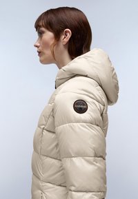 Chaqueta de plumas beige claro con capucha, que presenta secciones acolchadas y un parche de logo circular en el hombro izquierdo. Material suave y brillante.