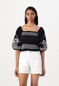 Svart topp med puff-erm, firkantet utringning, med hvite broderte mønstre og et samlet midjebånd, sammen med hvite shorts.