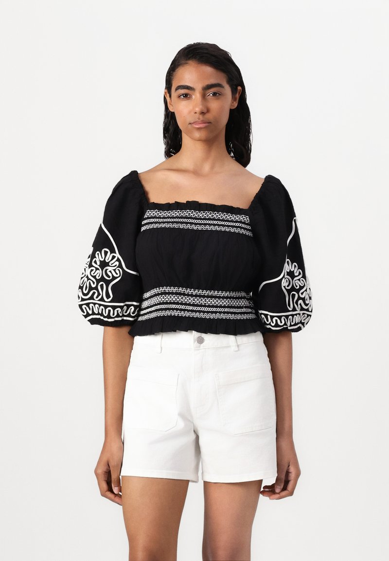 Svart topp med puff-erm, firkantet utringning, med hvite broderte mønstre og et samlet midjebånd, sammen med hvite shorts.