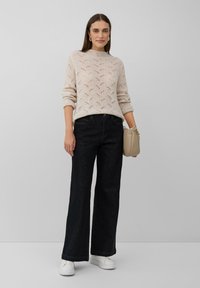 Femme debout portant un pull à motifs beige, un jean noir à jambes larges, des baskets blanches, des boucles d'oreilles en or et tenant un sac à main beige clair.