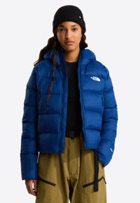 Chaqueta azul acolchada con capucha, con secciones acolchadas y un bolsillo con cremallera. Lleva puesta sobre una camiseta negra, combinada con pantalones kaki y un gorro negro.