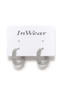Sudraba auskari ar riņķveida formu un teksturētu virsmu, rotāti ar caurspīdīgiem rhinestone akmeņiem, pasniegti uz baltas izstādes kartes ar "InWear" zīmolu.