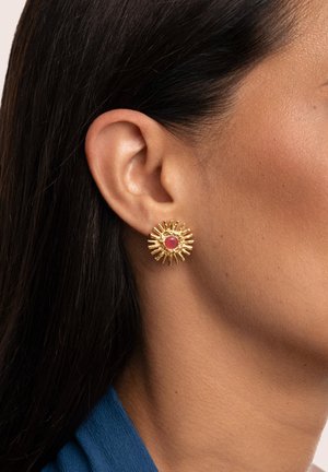 Orecchini a forma di sole in oro con una pietra rossa centrale. La superficie testurizzata con raggi che si estendono verso l'esterno aggiunge dimensione e interesse visivo.