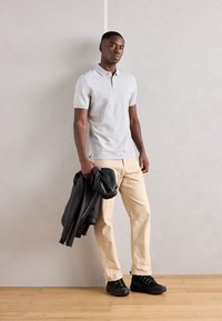 Grå polo t-shirt med korte ærmer, cremefarvede bukser, sorte sneakers og en sort læderjakke, der holdes i den ene hånd, stående på trægulv.