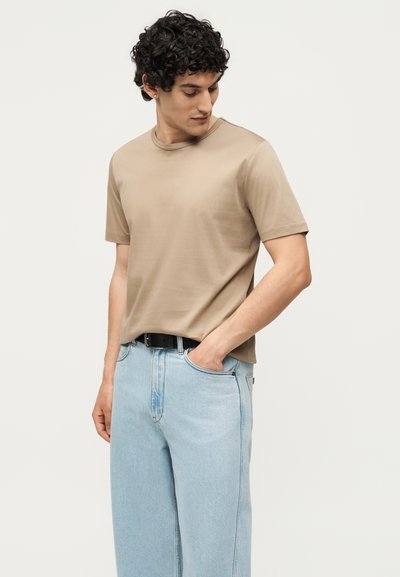 Άνδρας με μαύρα σγουρά μαλλιά που φοράει μπεζ κοντομάνικη μπλούζα μέσα σε ανοιχτόχρωμα μπλε τζιν με μαύρη ζώνη, κοιτάζει προς τα κάτω αριστερά.