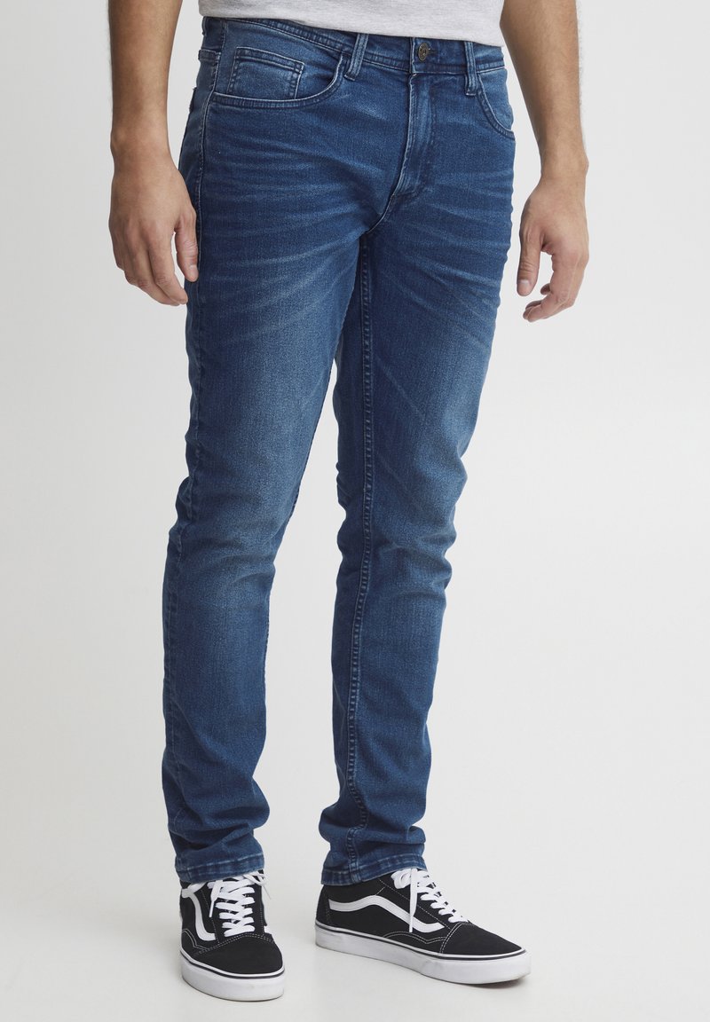 Jeans din denim albastru, skinny-fit, cu un model decolorat, cinci buzunare și închidere cu buton metalic, asortați cu adidași negri cu accente albe.