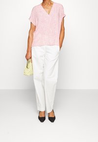 Helles rosa V-Ausschnitt-Oberteil mit kurzen Ärmeln, kombiniert mit weißen, weiten Hosen. Eine kleine geflochtene gelbe Handtasche und schwarze spitze Absatzschuhe runden den Look ab.