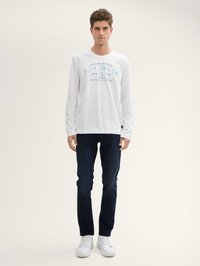 Camiseta blanca de manga larga con texto azul, jeans oscuros y zapatillas blancas. Diseño simple, cuello redondo, corte regular. Sin patrones ni acentos.