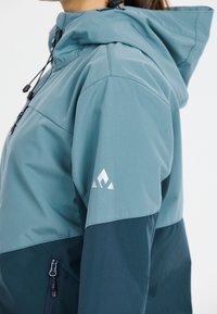 Lightweight, two-tone jacket in blue hues, featuring a hood, side zipper pocket, and logo accent on the sleeve. Smooth texture.
