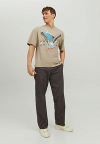 T-shirt beige con grafica di una farfalla in blu e nero, abbinato a pantaloni grigi scuri e sneakers bianche. Vestibilità casual e maniche corte.