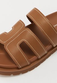 Brune lærsandaler med to brede stropper og hvide syninger, der har en formet fodseng og en struktureret gummisål for bedre greb.