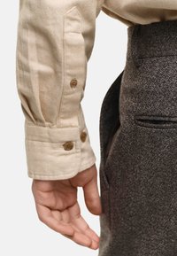 Puño de camisa en espiga beige con dos botones marrones, combinado con pantalones oscuros texturizados que presentan un detalle de bolsillo lateral.