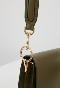 Sac à main en cuir vert olive avec fermoir en métal doré et anneau reliant une large bandoulière en cuir au sac.