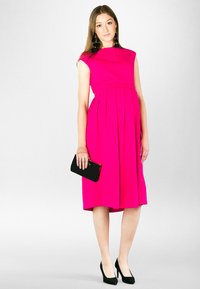 Fuksjowa sukienka w kształcie litery A z krótkimi rękawami i plisowanym dołem, w połączeniu z czarną kopertówką i czarnymi szpilkami, na jasnym tle.