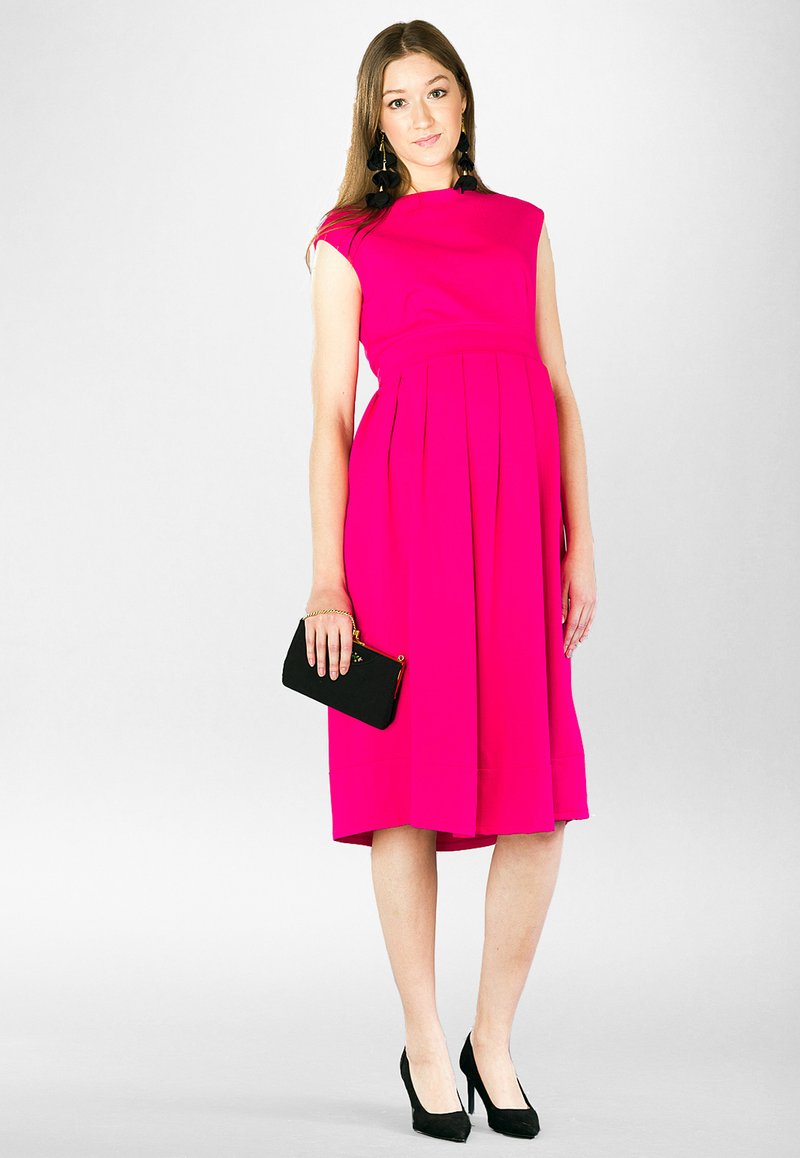 Fuksjowa sukienka w kształcie litery A z krótkimi rękawami i plisowanym dołem, w połączeniu z czarną kopertówką i czarnymi szpilkami, na jasnym tle.