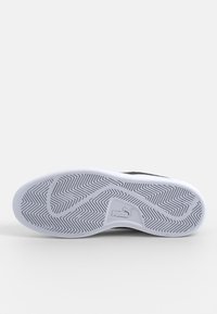 Semelle d’une basket Puma blanche et noire montrant un motif de crampons en zigzag et le logo Puma au centre sur un fond blanc uni.