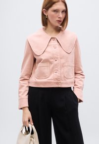 Ροζ cropped μπουφάν με μεγάλο γιακά, κουμπιά μπροστά και δύο μπροστινές τσέπες. Συνδυάζεται με μαύρο παντελόνι φαρδύ στο πόδι και κρατιέται με μπεζ τσάντα.