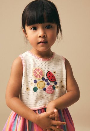 Ung pige med glat mørkt hår og pandehår iført en hvid ærmeløs top med farverig frugtbroderi og et flerfarvet skørt, hænderne foldet.