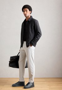 Man standing indoors wearing a dark jacket, light trousers, black sneakers, and holding a large black leather bag with one hand.