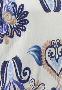 Tissu blanc avec des motifs floraux et en forme de cœur brodés en bleu et beige, présentant des volutes et des détails semblables à des feuilles.