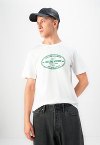 Ung mann som bærer svart caps, hvit grafisk T-skjorte og mørke jeans, ser mot venstre mot en enkel hvit bakgrunn.