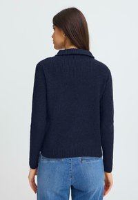 Hosszú, barna hajú nő, sötét kék bordás pulóverben és kék farmerben áll, háttal a nézőnek, egy világos, sima háttér előtt.