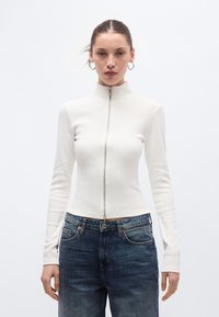 Γυναίκα με μαλλιά χτενισμένα προς τα πίσω, φορώντας ασημένια σκουλαρίκια κρίκους, λευκό μακρυμάνικο τοπ με φερμουάρ και μπλε τζιν, όρθια μπροστά σε απλό φόντο.