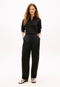 Kvinne med krøllete hår iført svart skjorte med knapper, vide svarte bukser og svarte flate sandaler med stropper, stående med hendene i lommene.