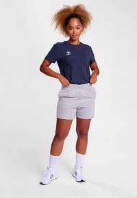 Marinblå kortärmad t-shirt med vitt tryck, kombinerad med grå shorts med elastisk midja. Modellen bär vita sneakers och ankelstrumpor.