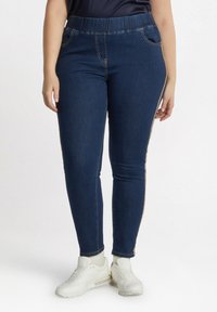 Leggings de mezclilla azul oscuro con cintura elástica, bolsillos laterales y costuras de acento dorado a lo largo de las costuras exteriores.