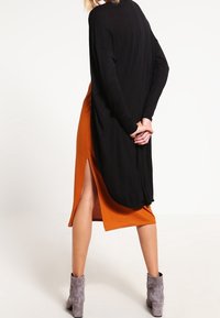 En person står med ryggen mot betraktaren, iklädd en svart lång kofta över en orange ribbad klänning, i kombination med grå ankelstövlar.