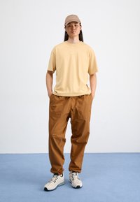 Beige rövid ujjú póló logóval, barna cargo nadrág és krémszínű tornacipő. Világosbarna sapkával kiegészítve. Egyszerű, sportos stílus.