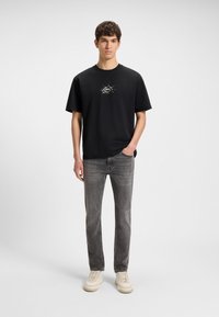 Fiatal férfi áll egy kézzel a zsebében, fekete „BOSS” logós pólót, szürke farmert és fehér tornacipőt visel, egyszínű háttér előtt.