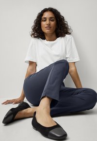 T-shirt bianco, pantaloni navy con spacco laterale e ballerine in suede nere con dettaglio grigio sulla punta. Il modello è seduto con una gamba incrociata.