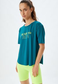 Teal træningstrøje med teksten "READY TO RUN" i gul print. Korte ærmer, rund halsudskæring, parret med neongrøn shorts. Glat stoftekstur.
