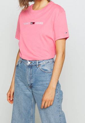 Kobieta ubraną w różowy t-shirt Tommy Jeans z wyszywanym logo oraz jasnoniebieskie dżinsy z wysokim stanem, stojąca na jednolitym tle.