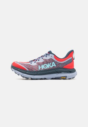 Hellgrauer und roter Laufschuh mit schwarzer Sohle, blauem "HOKA"-Logo und Vibram-Profil, von der Seite auf weißem Hintergrund gezeigt.