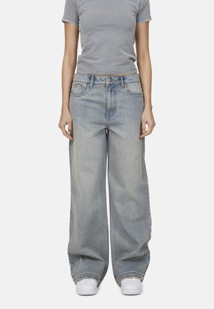 Jean large en denim bleu clair avec une taille haute, cinq poches et des détails de couture prononcés, associés à des baskets blanches.