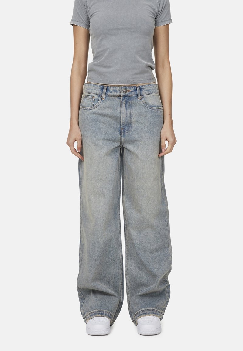 Jean large en denim bleu clair avec une taille haute, cinq poches et des détails de couture prononcés, associés à des baskets blanches.