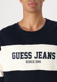 Marineblauwe crewneck sweatshirt met een crèmekleurige streep. Dikke "GUESS JEANS" logo en "SINCE 1981" in donkere letters. Katoenen stof met een gladde textuur.